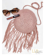 A round, crocheted purse with fringe hanging down, a pair of sunglasses, and a tube of lip gloss are arranged on a white background.