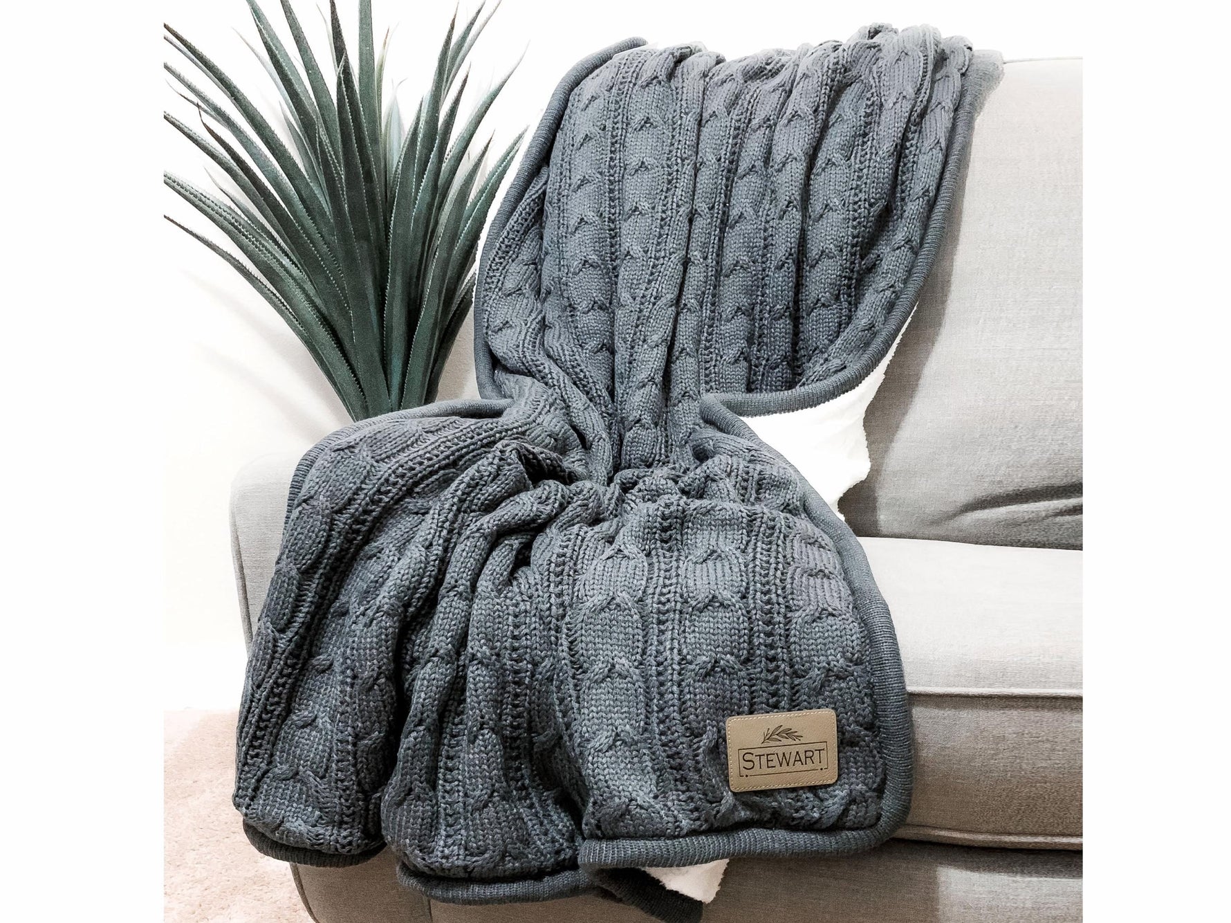 A gray knitted throw blanket is draped over the back of a gray sofa, with a small label on the blanket.